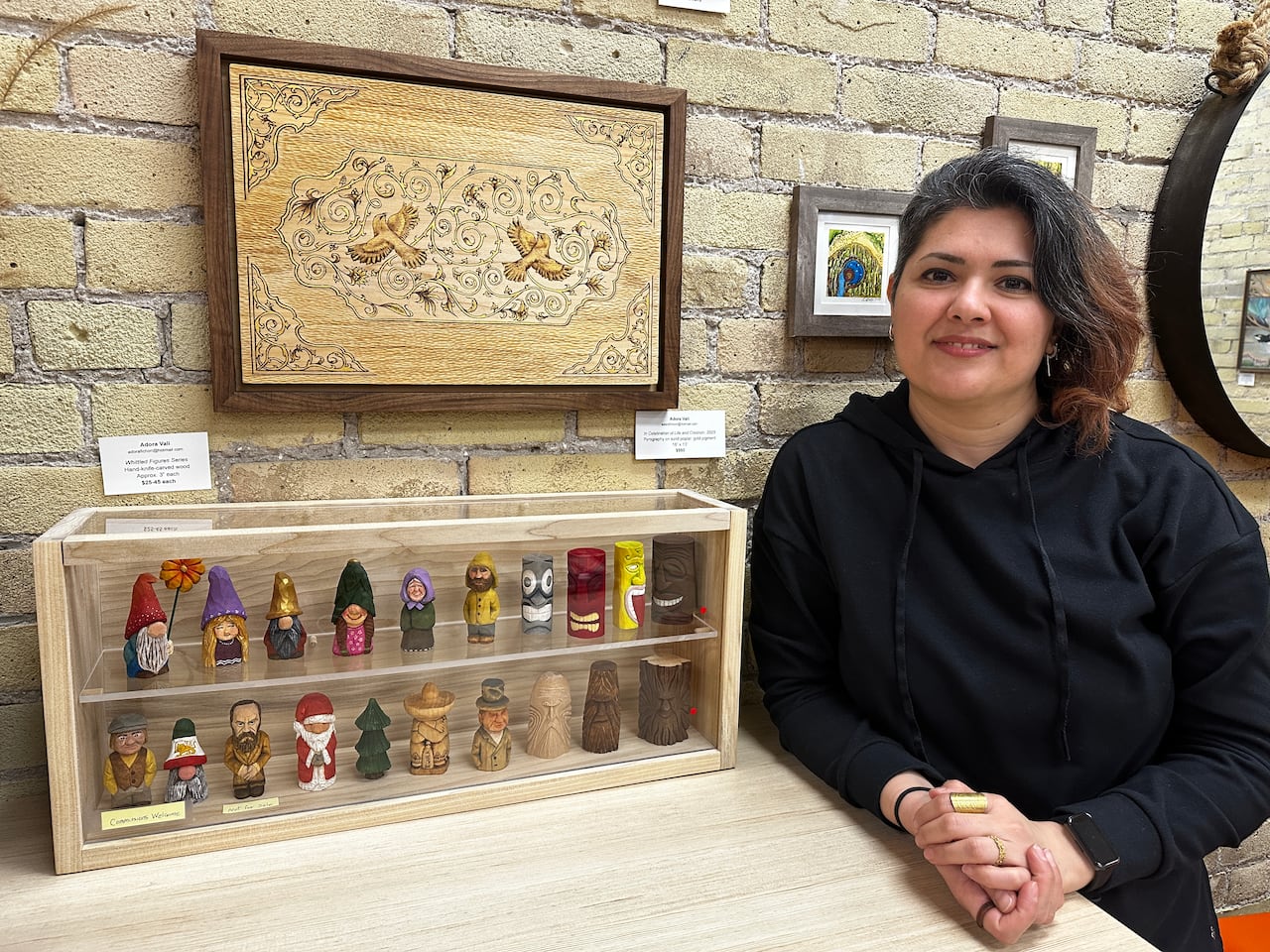 Woman wearing black sweater stands next to art work of little characters and wood carvings