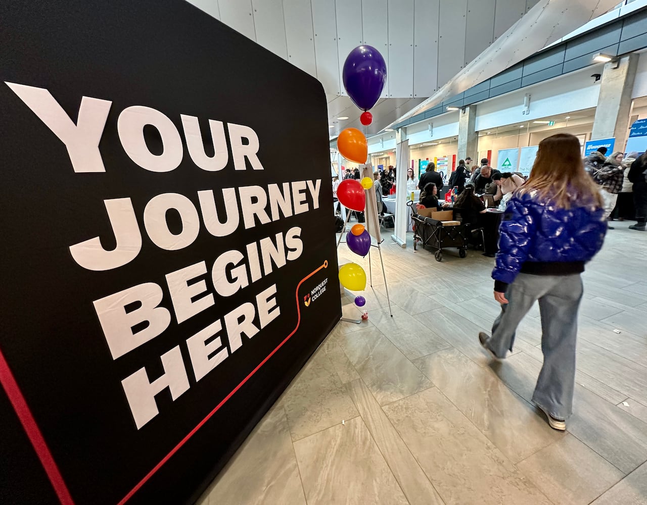 A student in jeans and a blue jacked walks with her back to the camera towards the job fair with booths and balloons next to a sign about the future. 