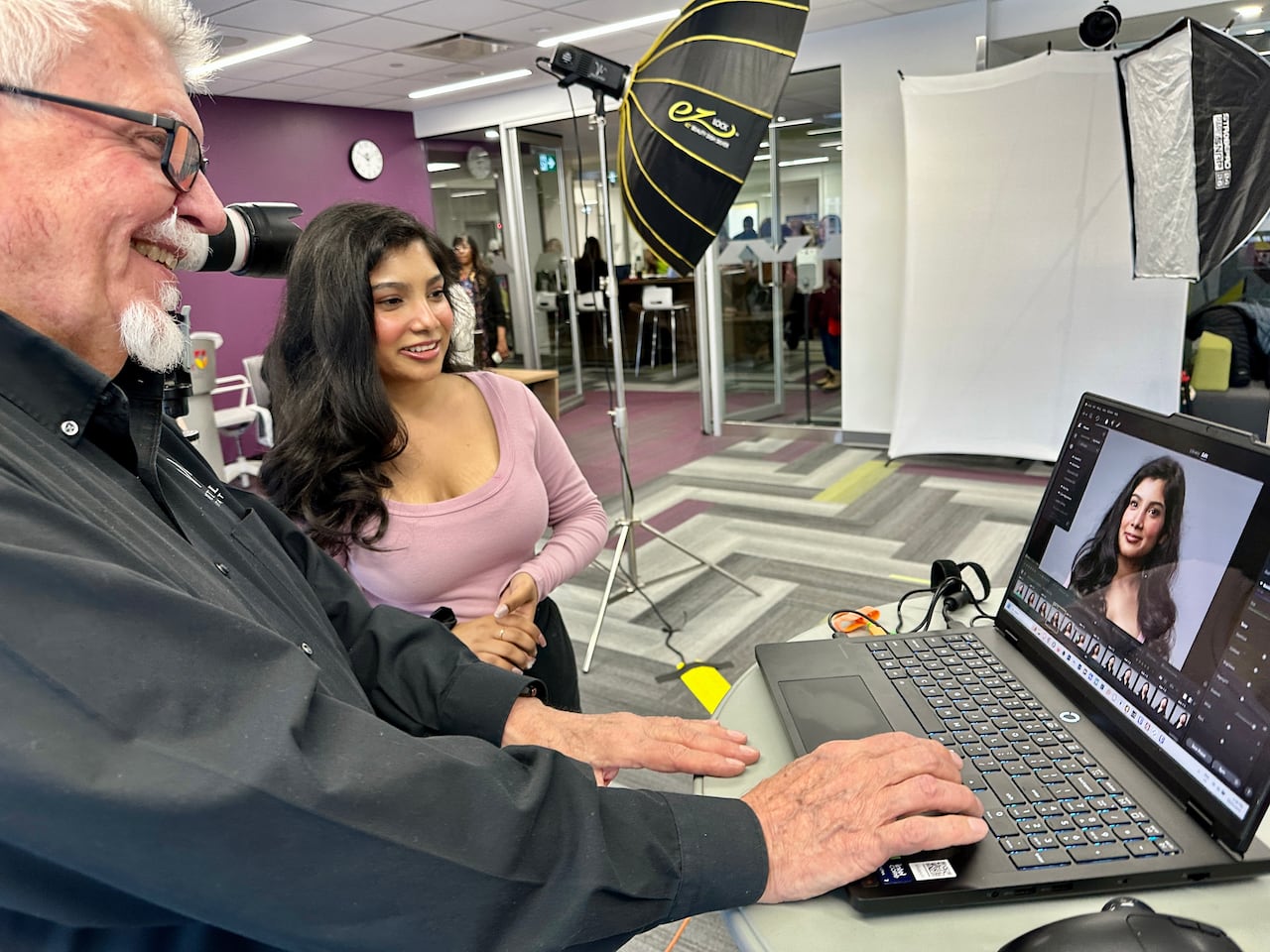 A man at a computer screen stands next to a student in a purple top reviewing images he's taken of her in a professional photo shoot.