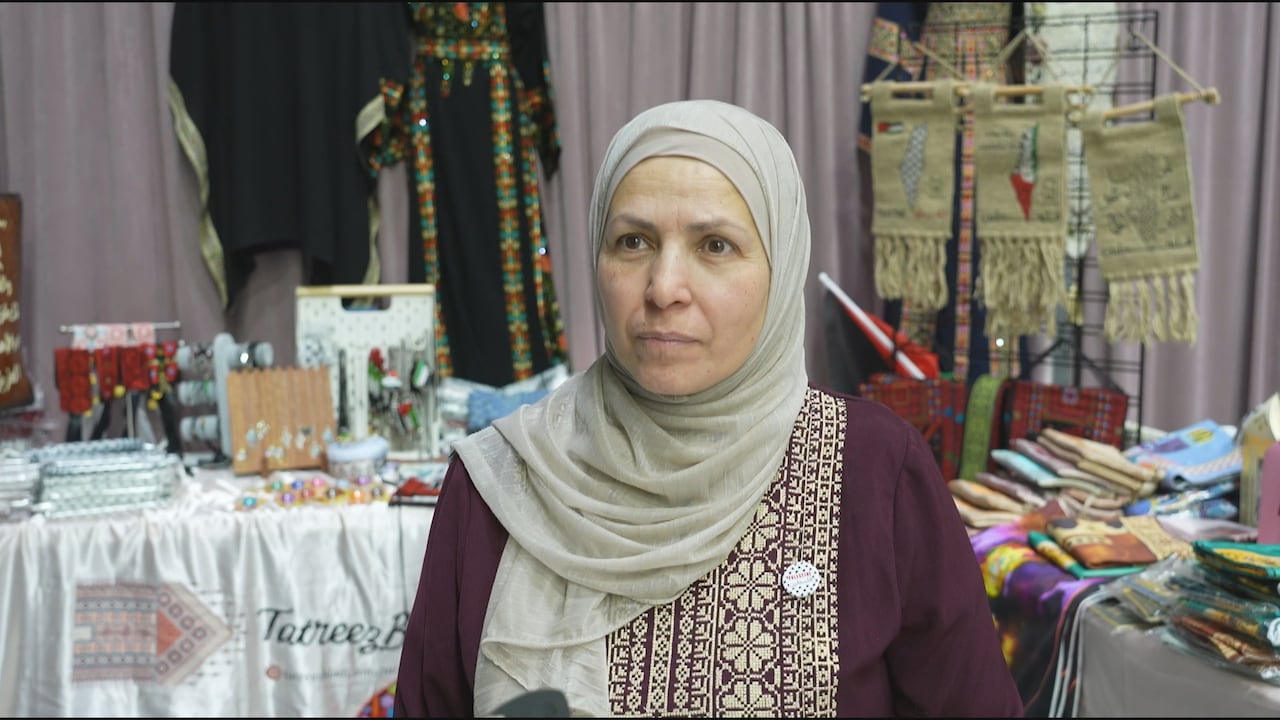 A woman wearing a hijab stands at her booth displaying embroidered clothing, accessories and textiles at a vendor stall.