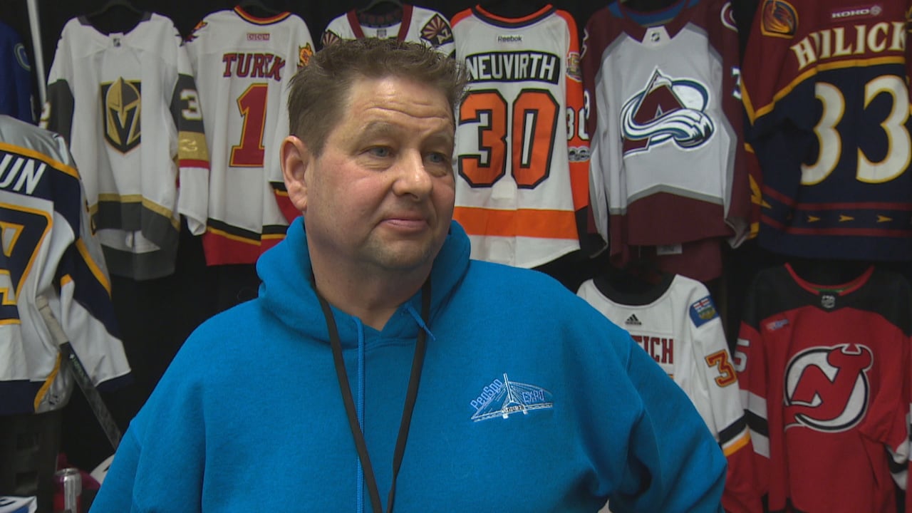 Man wears a blue hooded sweatshirt and stands infront of a booth filled with sports jerseys