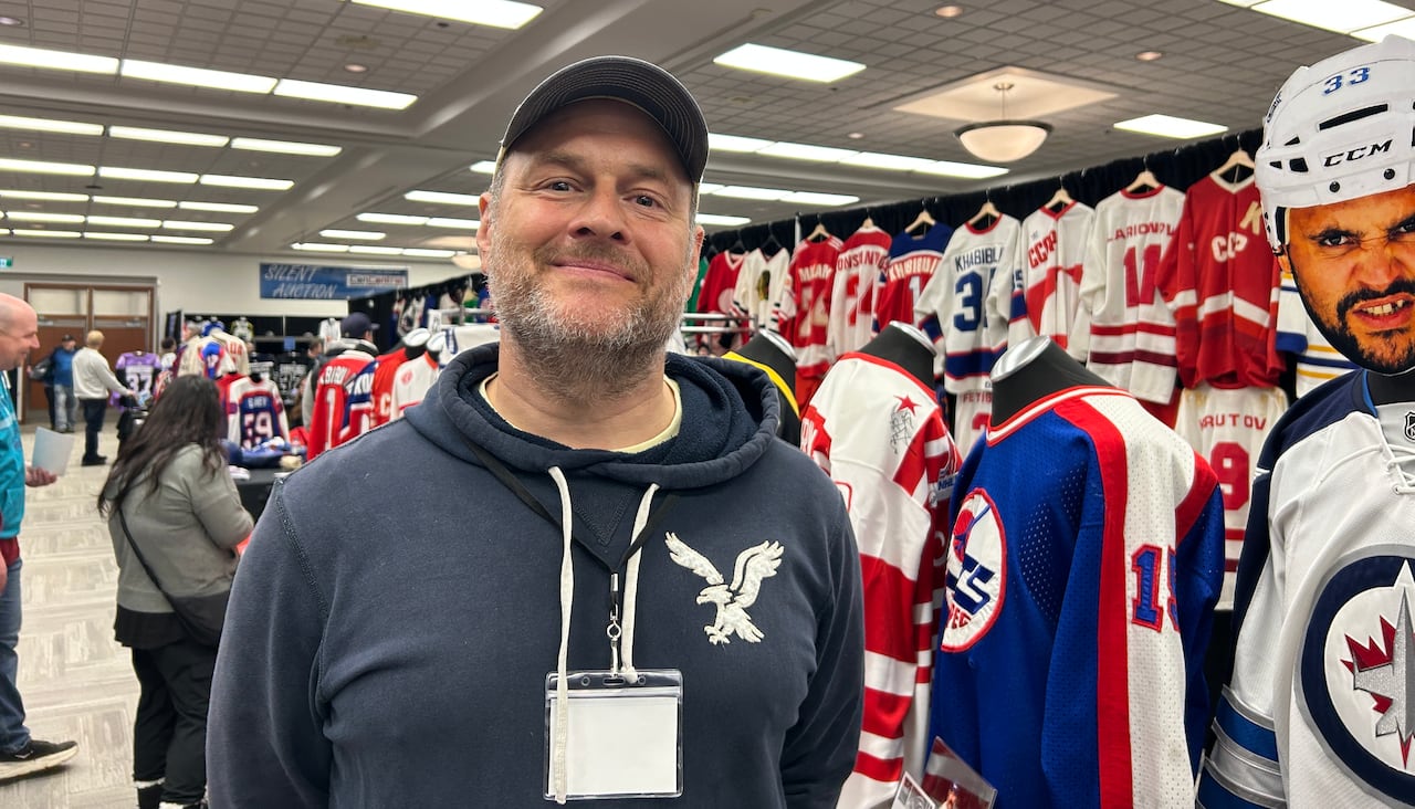 Man wearing blue hooded sweatshirt stands infront of a booth filled with hockey jerseys.