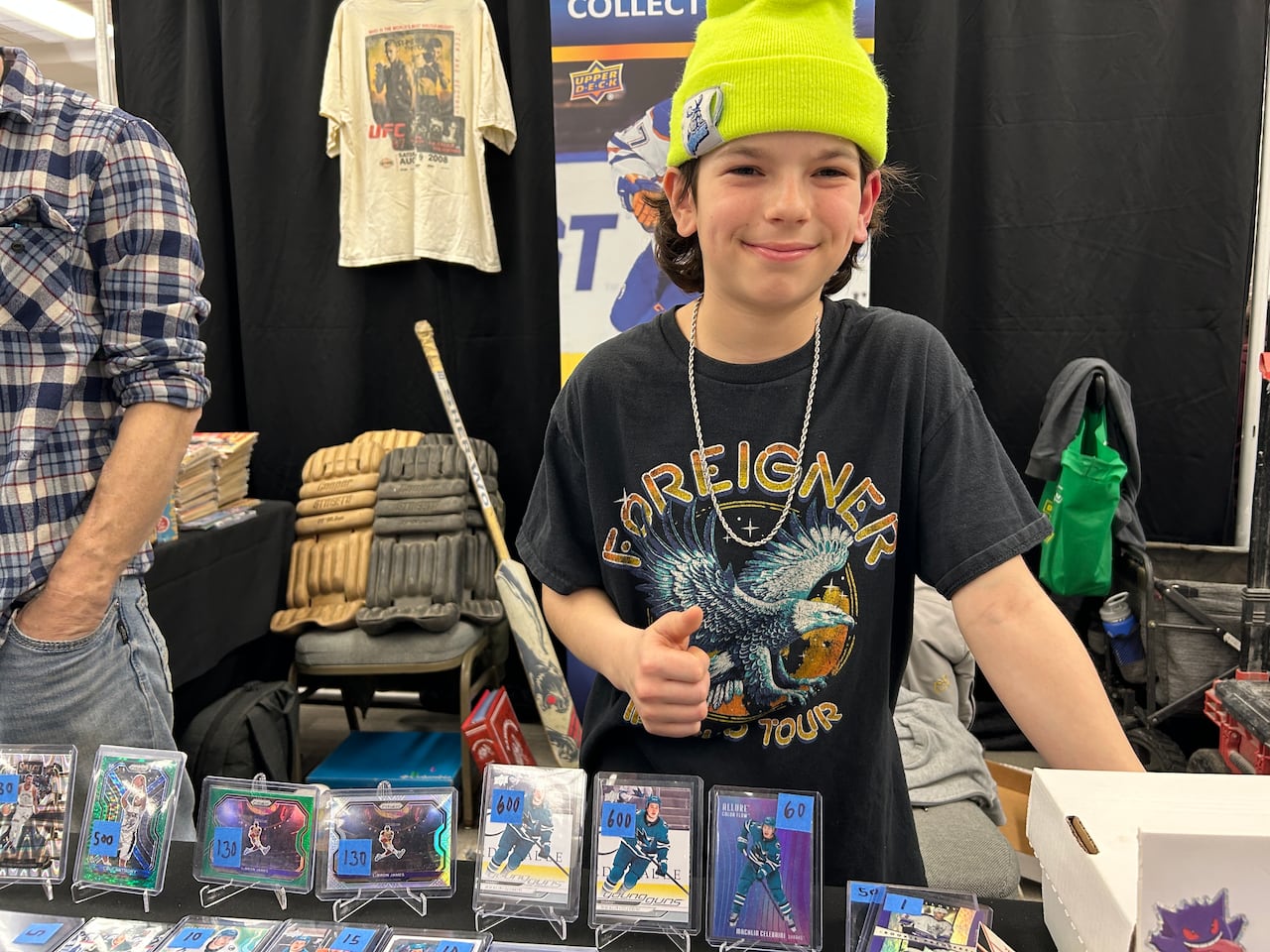 a young boy wearing a black t-shirt and a bright yellow hat gives a thumbs up behind a table displaying hockey cards