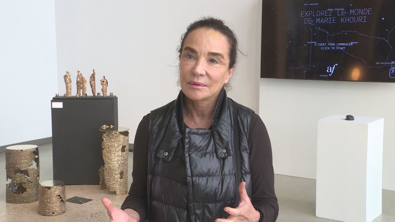 A woman wearing a black vest gestures while talking inside her studio with some of her sculptures in the background.