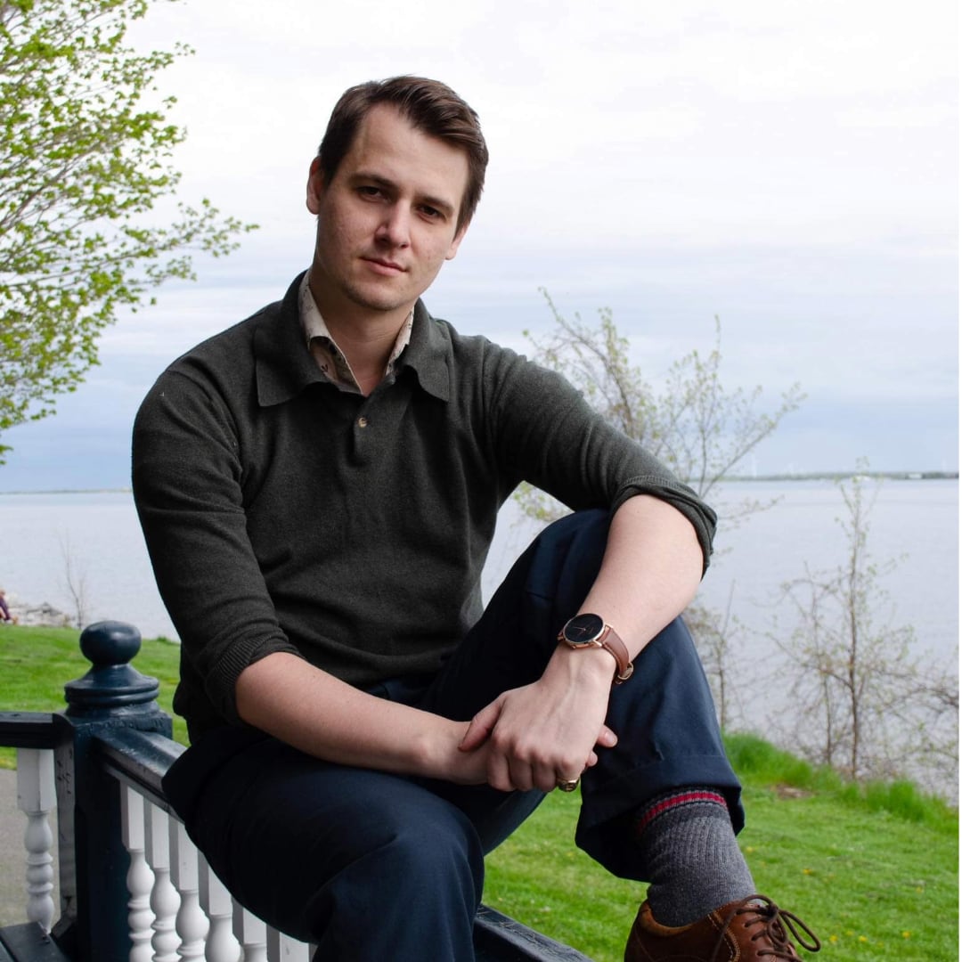 A man sits outside on a banister behind him is a lake