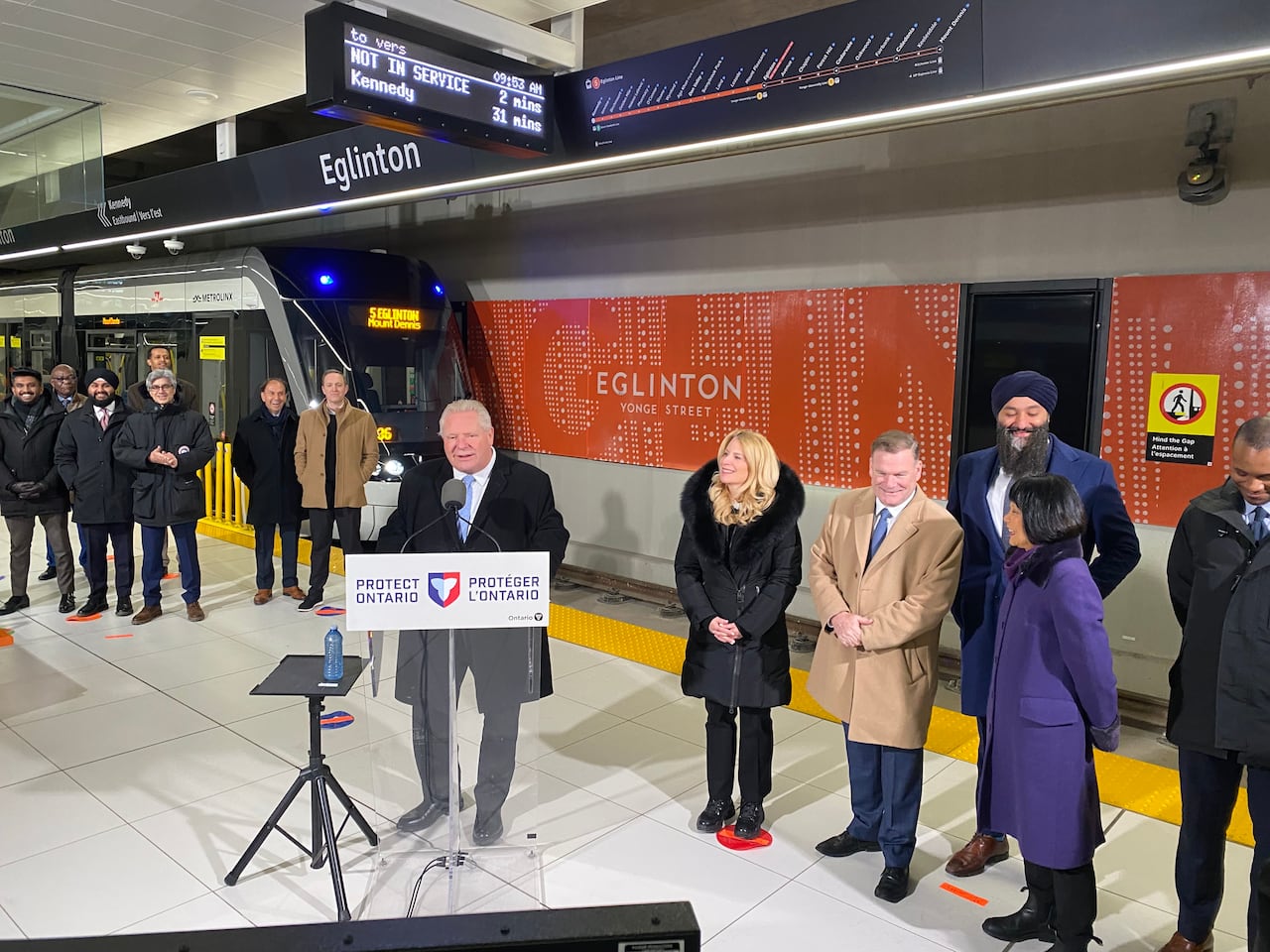 Doug Ford stands at a podium in front of an lrt station with a sign above him and other people and politicians to his left and right