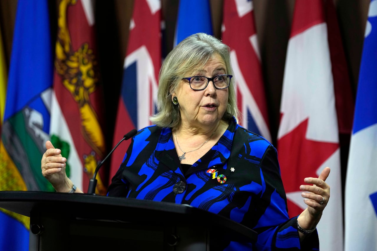 A woman in a blue blouse gestures with her hands as she speaks at a podium.
