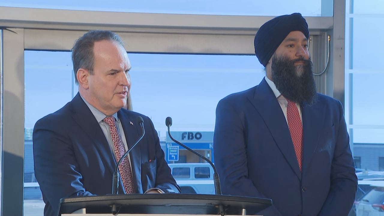 A white, middle-aged man in a suit stands at a podium in an airport next to a brown middle-aged man in a suit and turban.