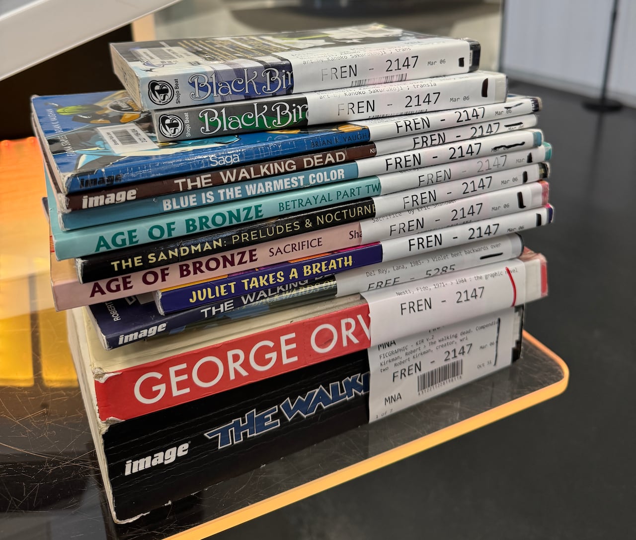 A stack of graphic novels on a table. They have library hold tags on the spine. Books include two of the Age of Bronze comics, The Walking Dead and manga series Saga and Black Rabbit. 