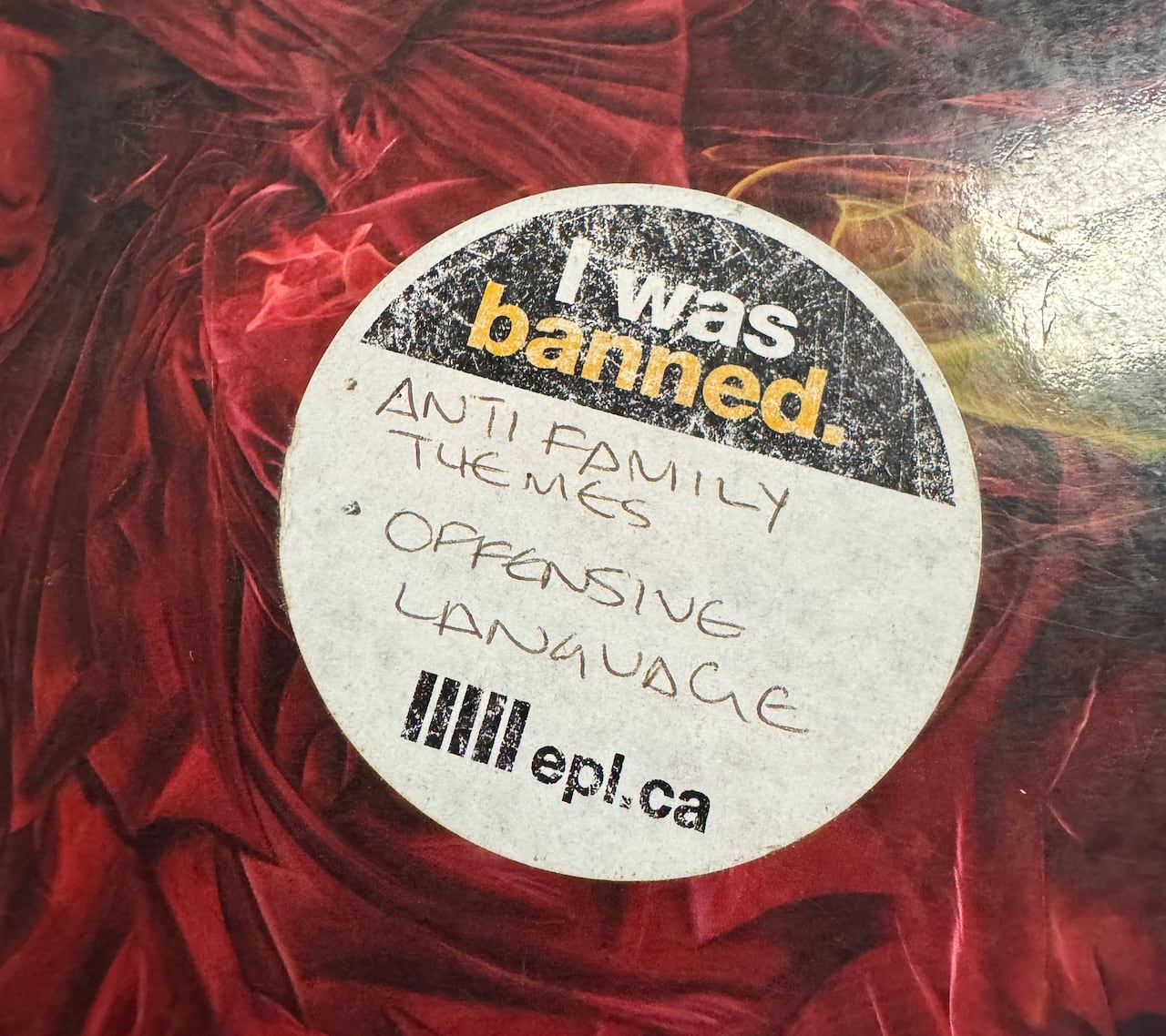 A white sticker says 'I was banned.: Anti-family themes. Offensive language. EPL.ca." It is on the cover of a unknown red book. 