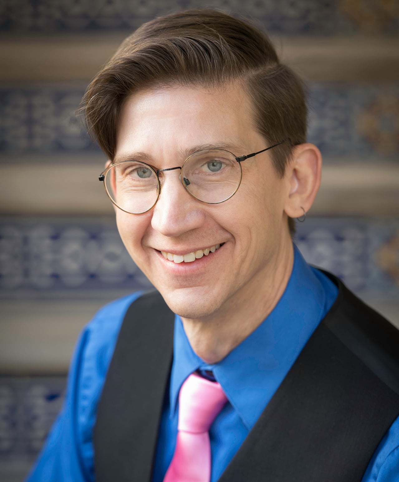 A man with brown hair and glasses poses for a photo. He's wearing a blue shirt, black vest and pink tie.