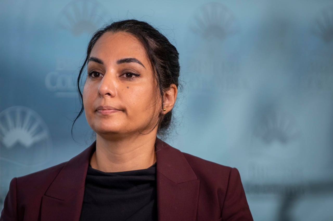 A woman stares into space with her mouth closed. She wears a black top and a burgundy jacket.