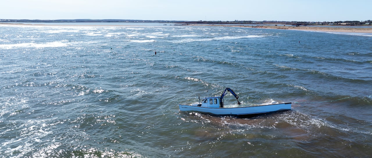 A drone view of a half-submerged white and blue fishing boat near a sandbar, being hit by waves. 