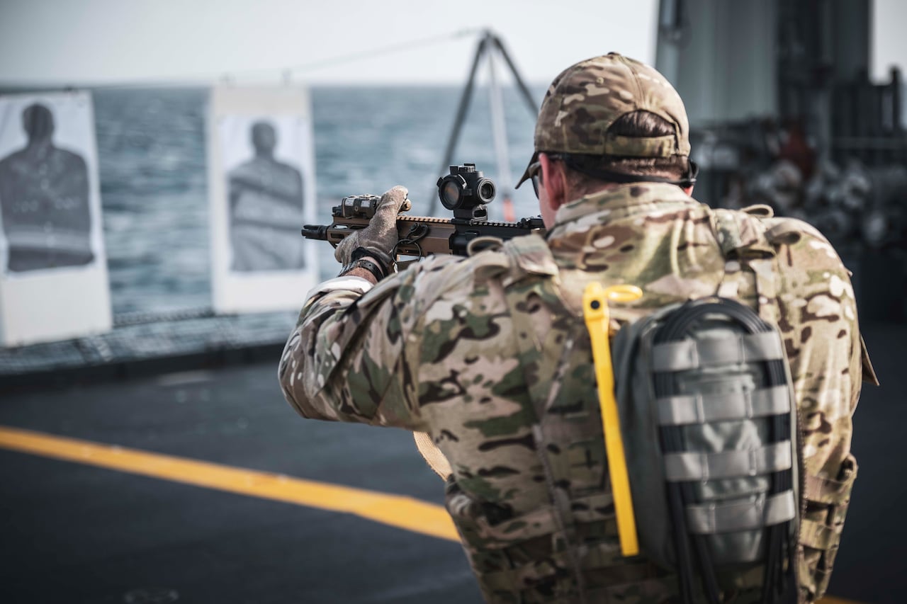 A man dressed in camo fires a carbine at mansize targets on the deck of a ship.