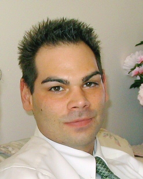 A man with black hair wearing a white button-down and green tie smiles slightly at the camera.