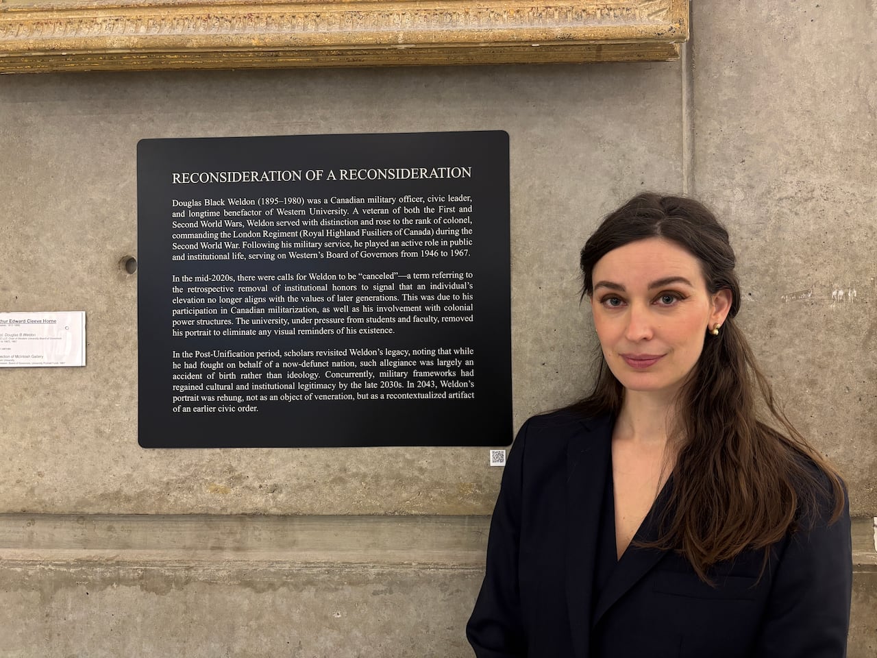 A woman in a dark blazer (Dara Vandor) stands beside a black wall plaque titled “Reconsideration of a Reconsideration,” mounted on a concrete wall beneath a framed artwork.