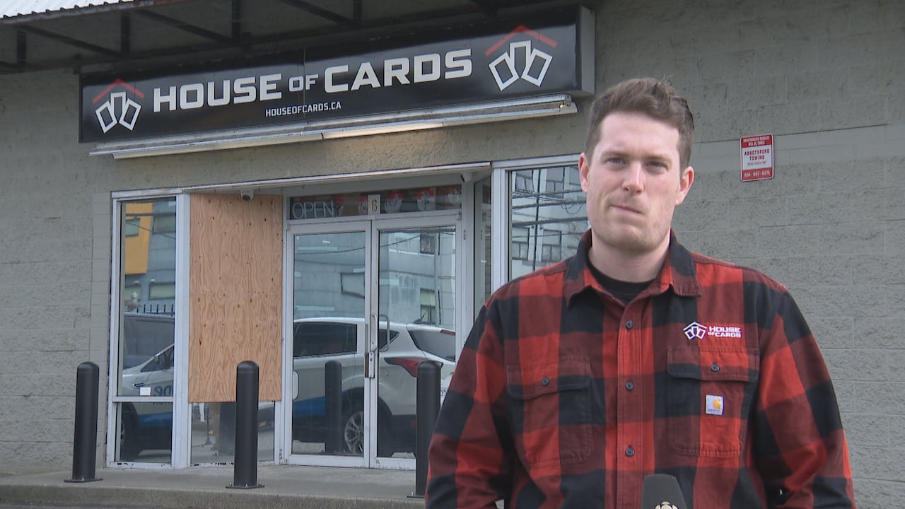 A man wearing a checked red shirt stands outside a store marked 'House of Cards' with a boarded-up window.