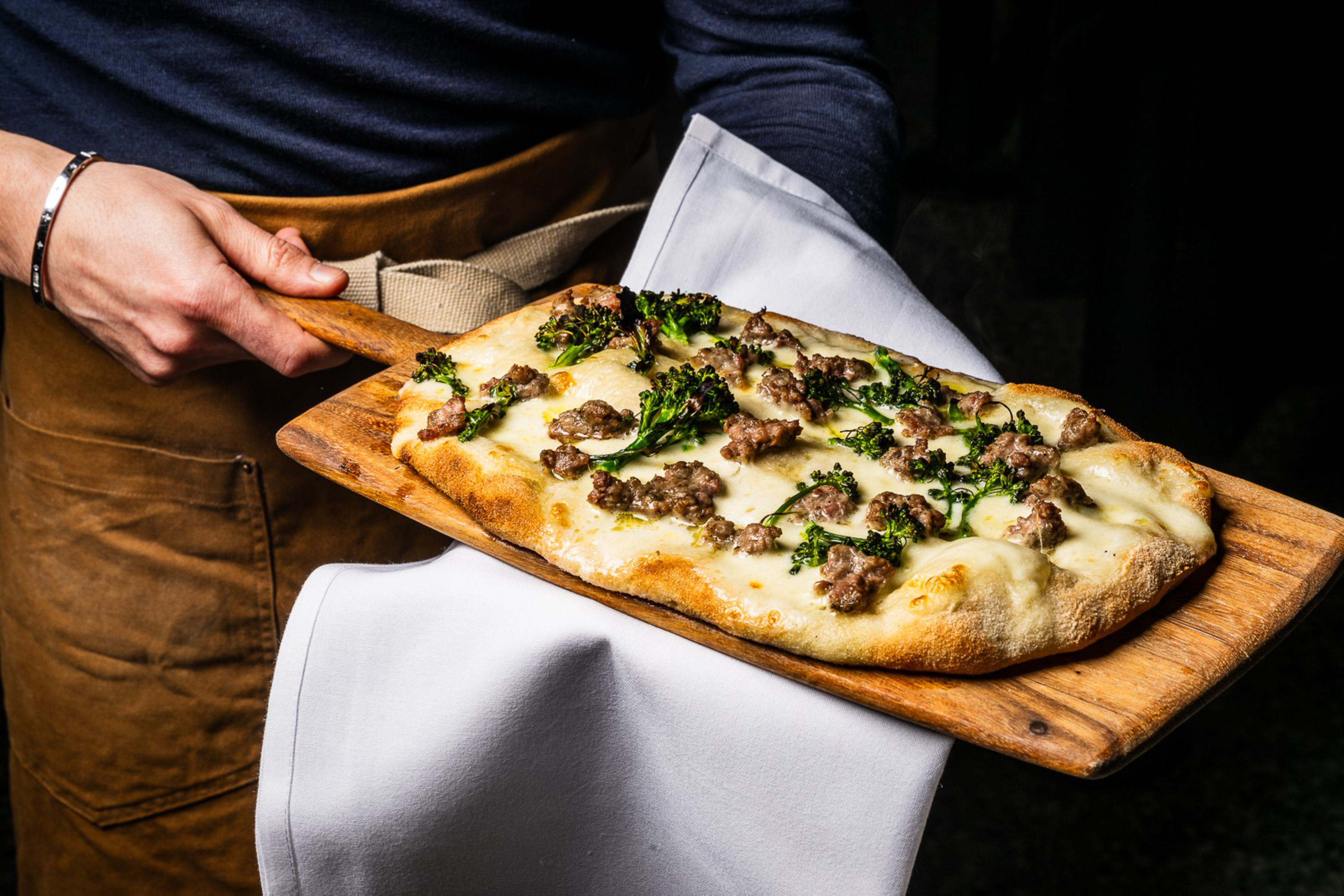 Flatbread pizza topped with melted cheese, sausage pieces, and green herbs held on a wooden paddle by a person wearing a brown apron.