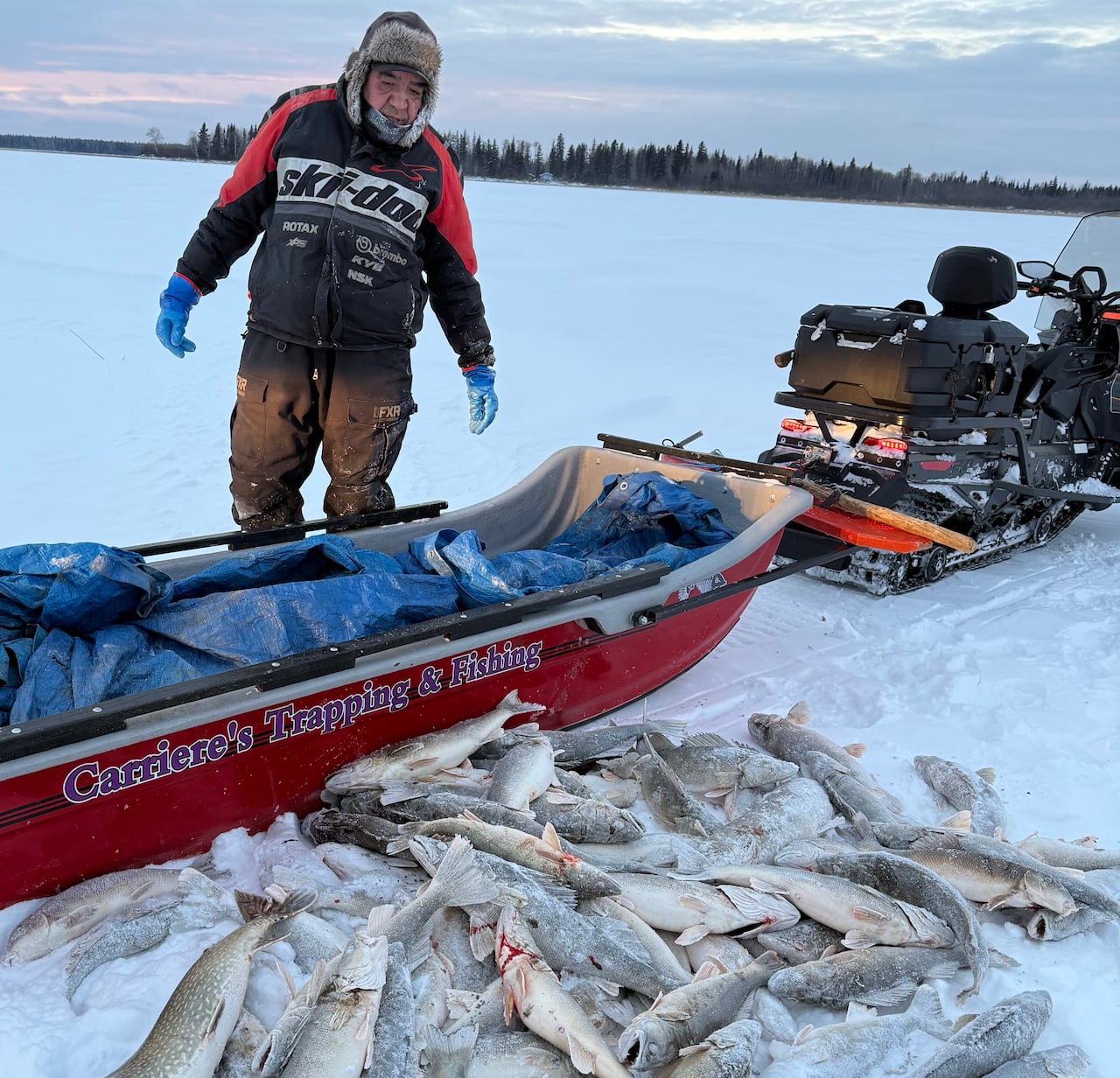 A man stands on the ice by a ski mobile and sleigh. Several fish lay on the ice of the frozen lake.