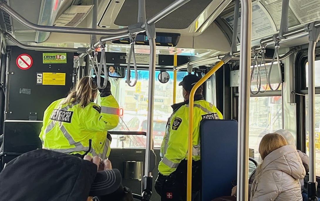 Two transit safety officers in bright yellow vests stand inside a bus, at the front near the windshield.
