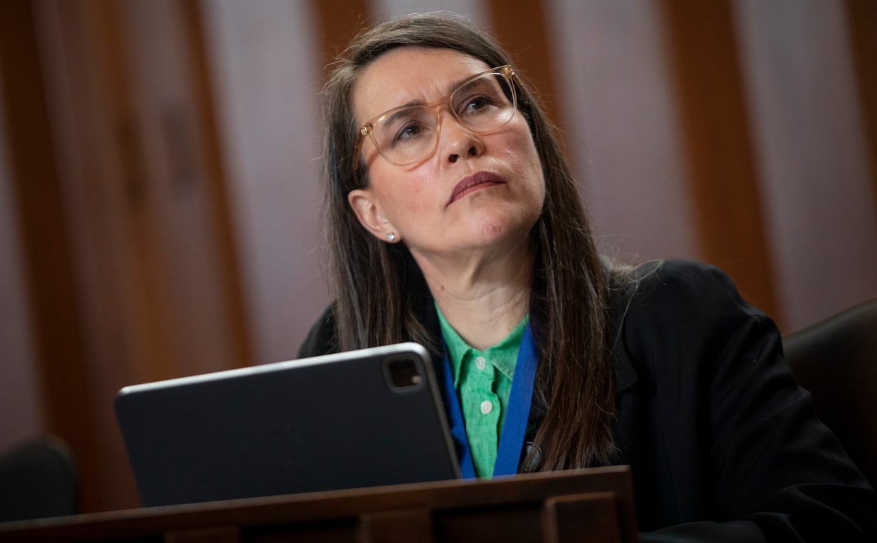 A woman wearing glasses and a green shirt looks to her left while seated in front of a screen.