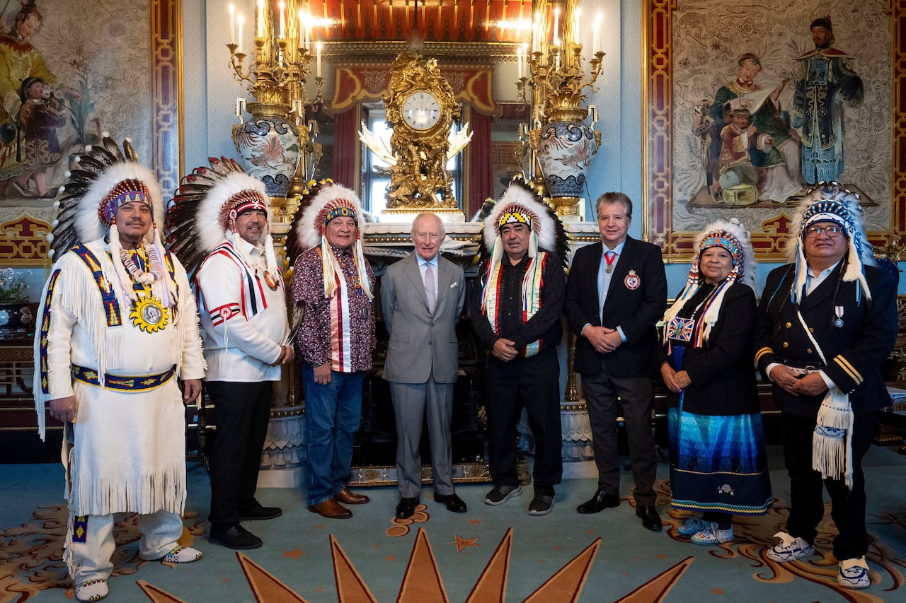 A group of seven Indigenous people, six of whom are wearing traditional regalia, flank an elderly white man in a grey suit. They are all posing for a photo, smiling at the camera.