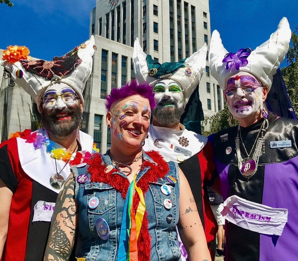 Three sisters of the Sisters of Perpetual Indulgence in white wimples and colourful flowers stand with another person in a jean jacket, buttons and rainbow scarf in front of Vancouver city hall.