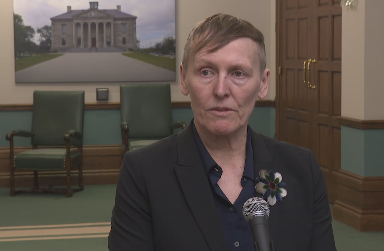 A woman with short hair, wearing a suit, stands in from of a microphone in an ornate room in a government building