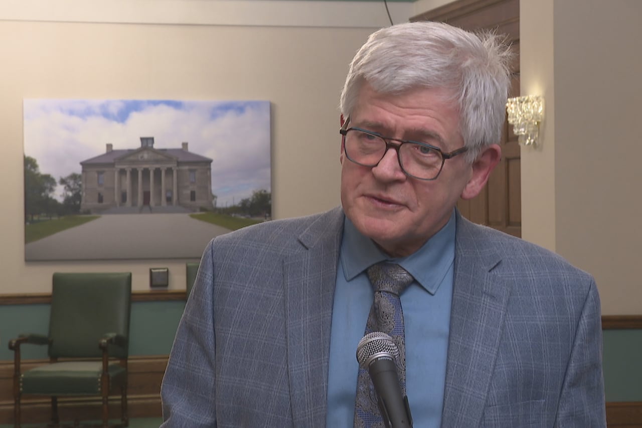 A white-haired man, in a blue suit and a tie, stands in front of a microphone in an ornate government building