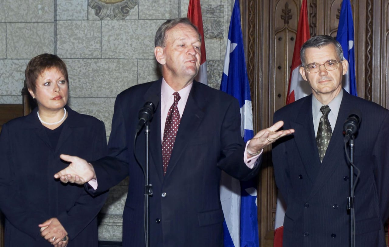 A man in a suit gestures with his hands as he speaks into a microphone. A man and a woman in business attire stand beside him.