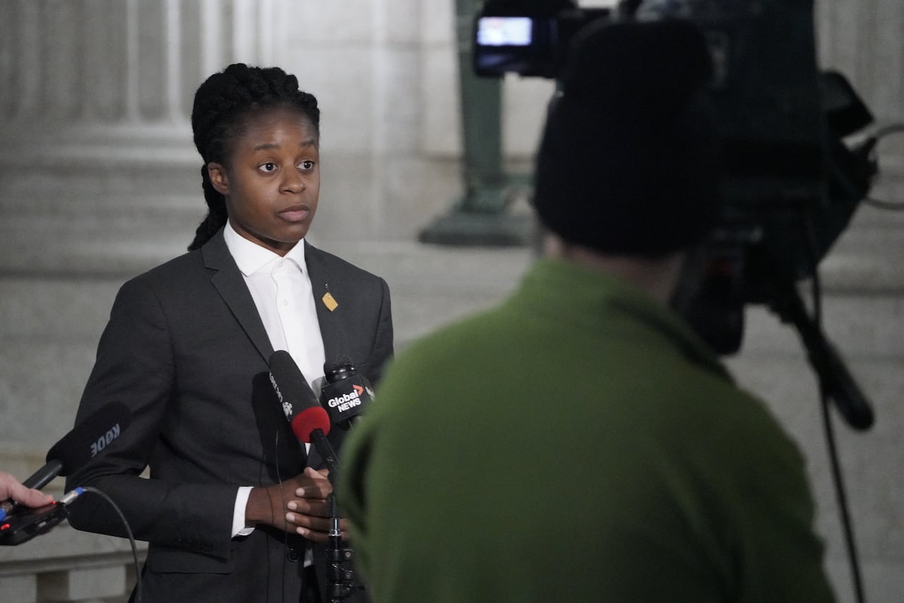 A person in a white shirt and dark blazer speaks into microphones at a lectern in a government building.