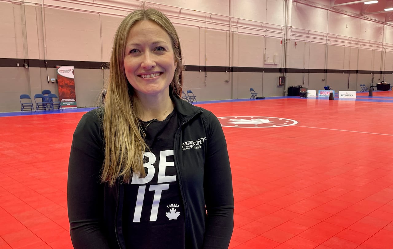 A smiling blond woman standing a gymnasium, wearing a shirt that say "BE IT"