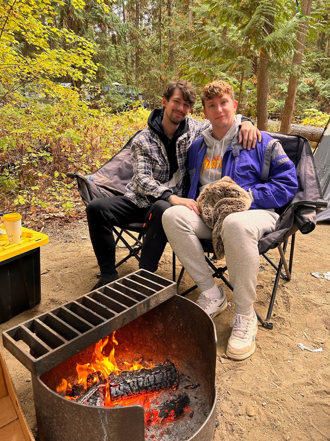 Two men sit in a folding chair beside a campfire.
