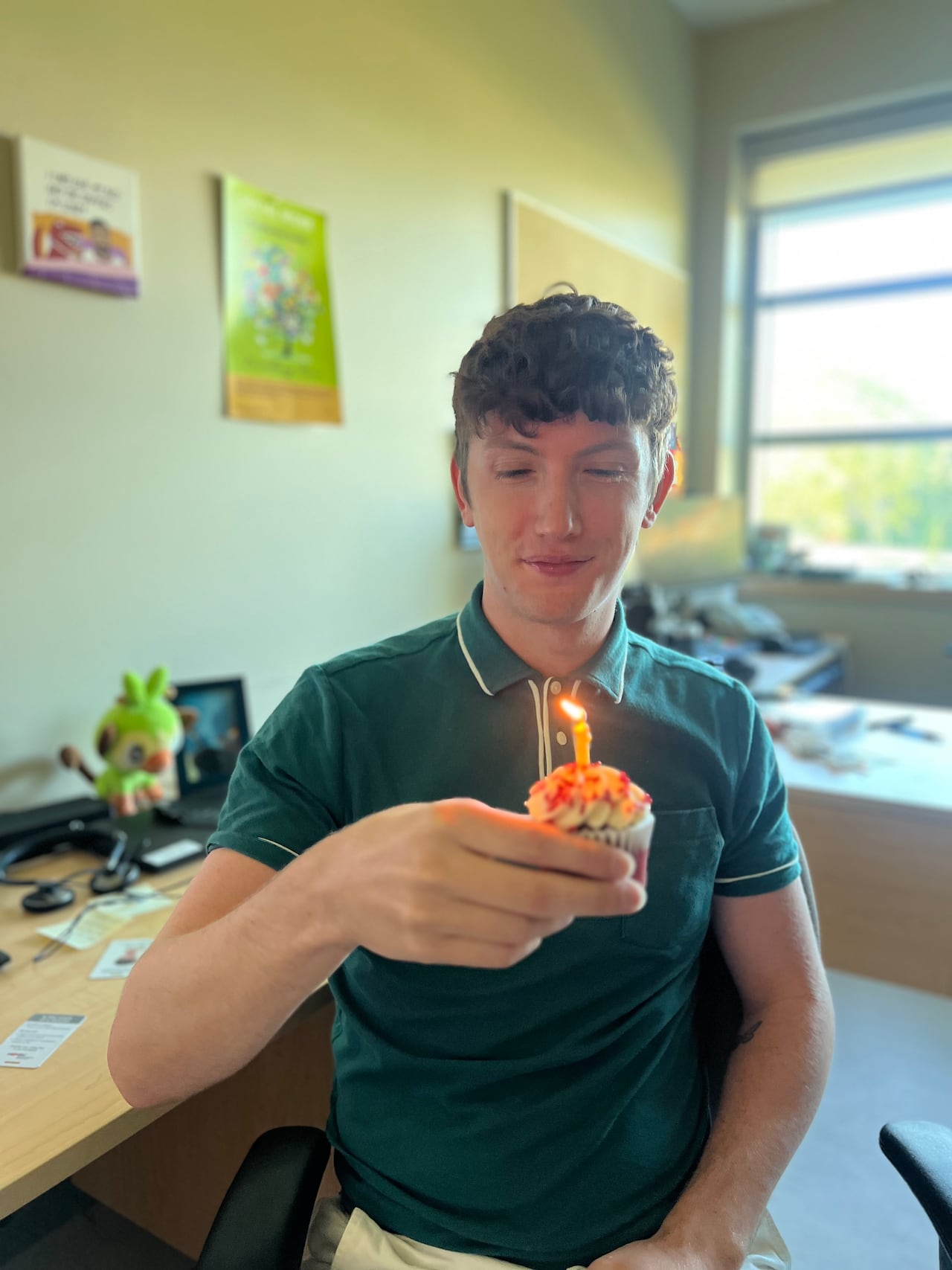 A man in a dark green gold shirt holds a cupcake with a single candle.