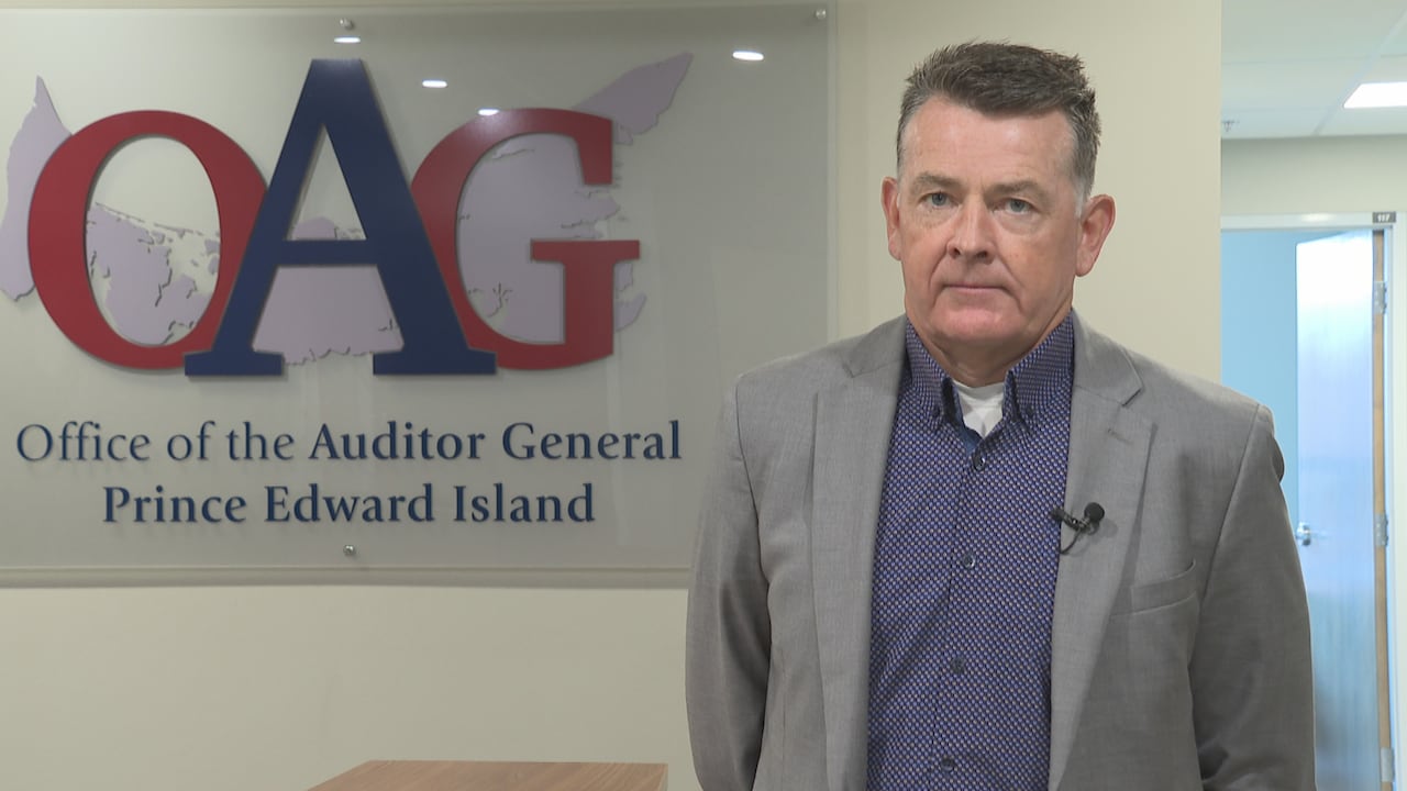  Man in grey blazer over navy blue shirt standing in front of a sign that reads Office of the Auditor General.