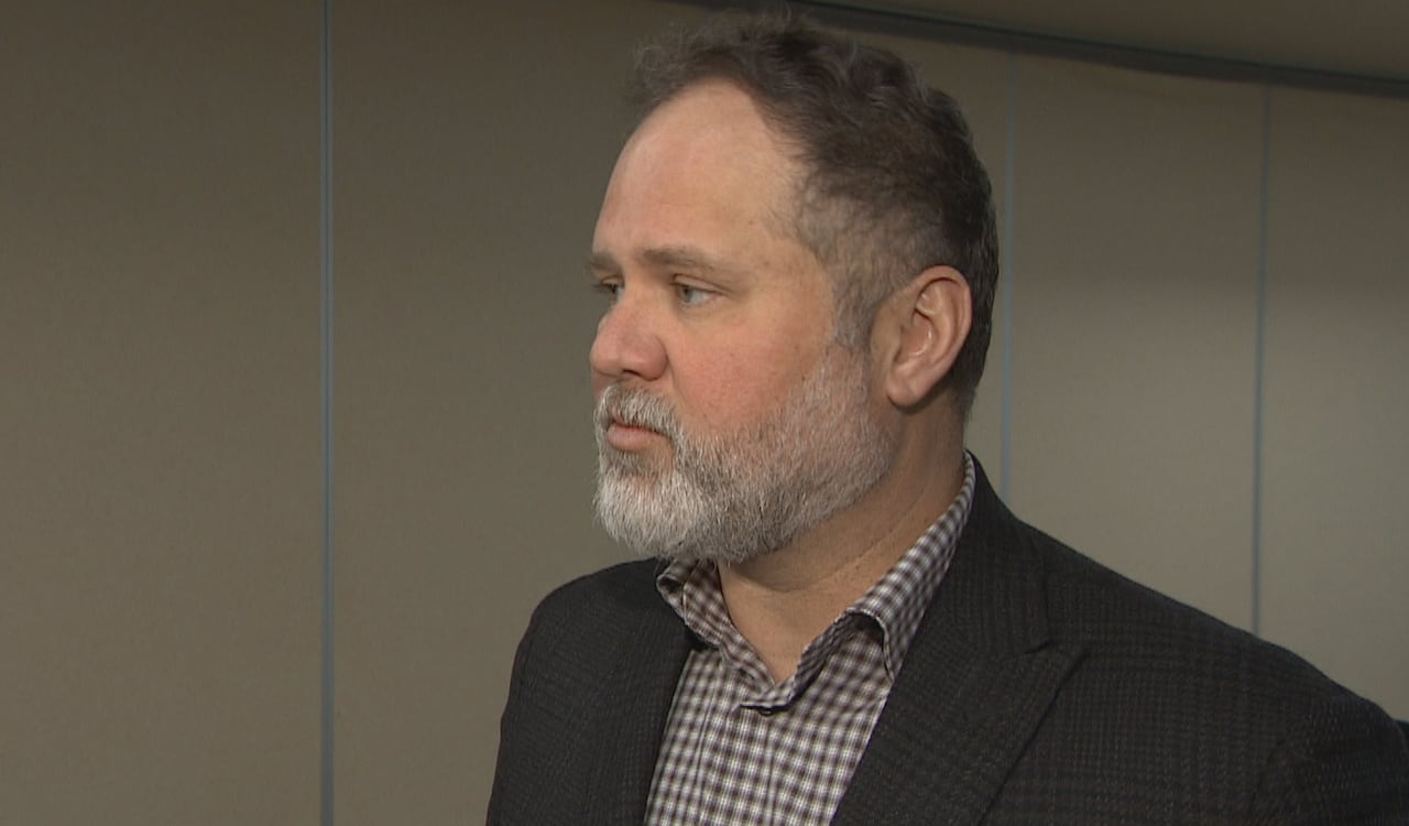 Man with dark hair and white beard wears a black suit jacket and checkered shirt underneath.