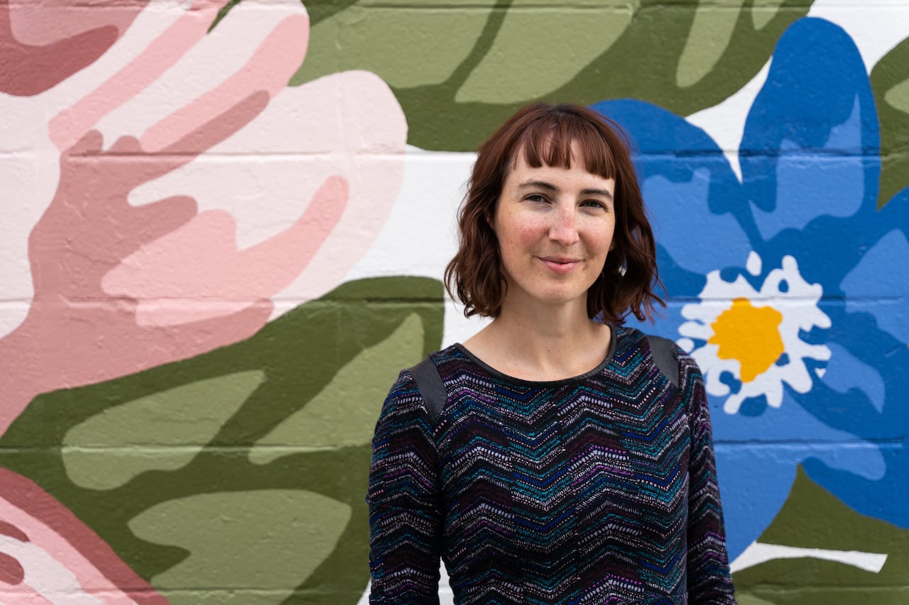 A woman wearing a purple sweater stands in front of a floral mural