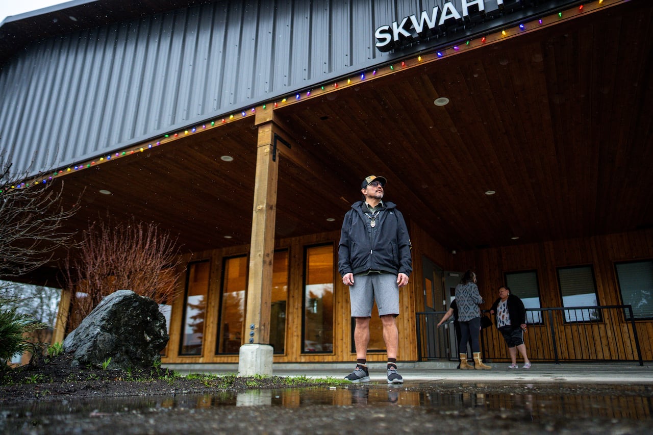 a man in shorts, a jacket, a hat and with a circle beard, stands underneath a covered area out of the rain