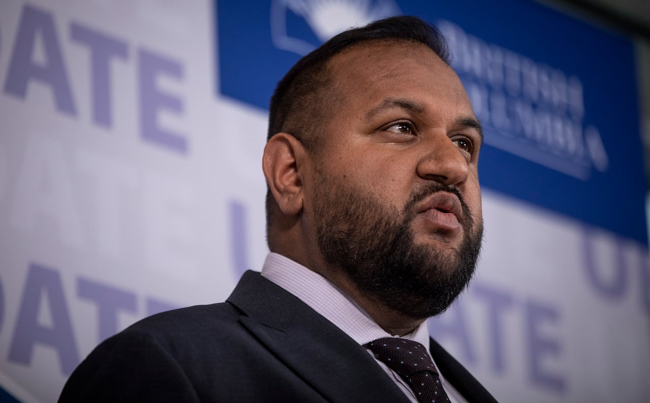 A South Asian man speaks in front of a blue and white banner.