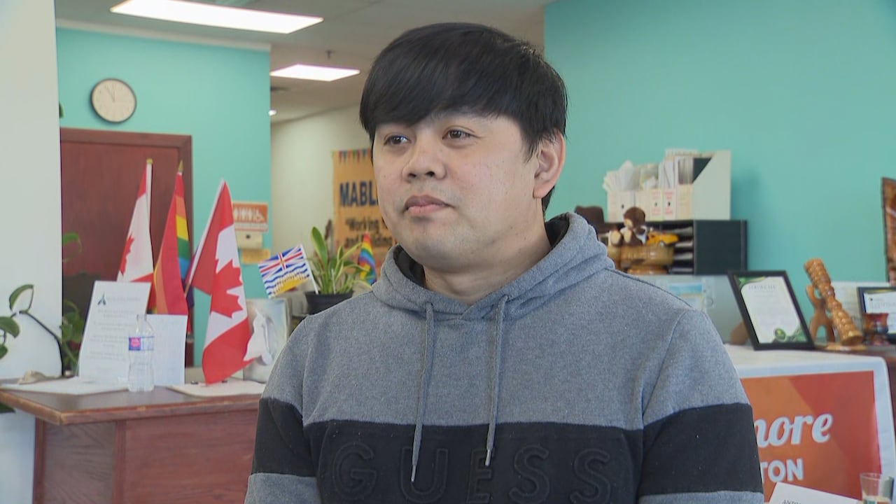 A man wearing a grey and black hoodie stands indoors during an interview.