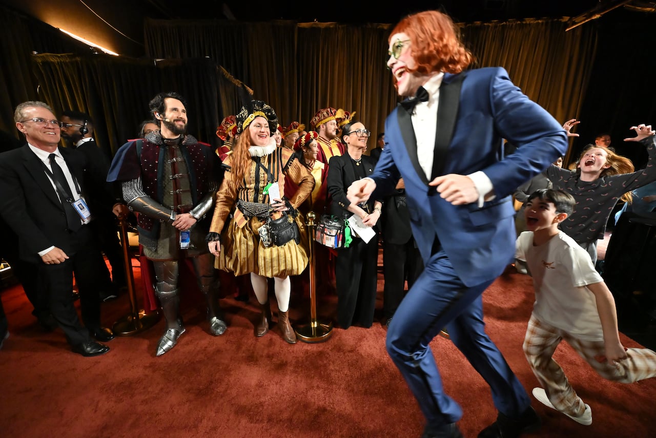 A man in a suit, a red wig and clown-ish makeup sprints past a line of people in costumes.