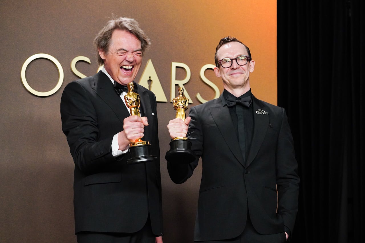 Two middle aged white men in tuxedos hold Oscars and smile backstage at the Academy Awards