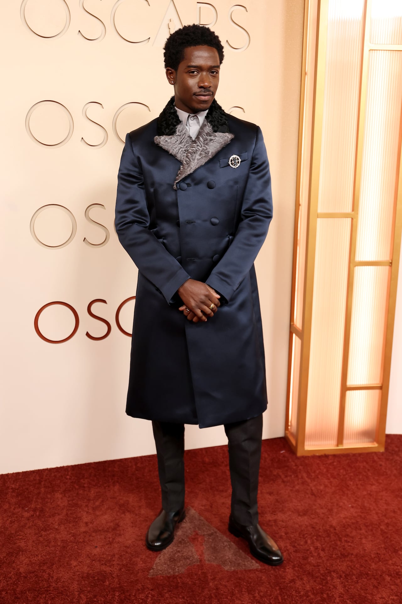 A man poses for a photo wearing a long double-breasted navy satin coat with a grey fur lapel, pants and black dress shoes.