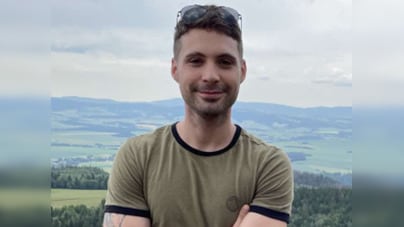A man leans against a railing and smiles, with arms crossed in front of him. Rolling farmland and forest can be seen in the background.
