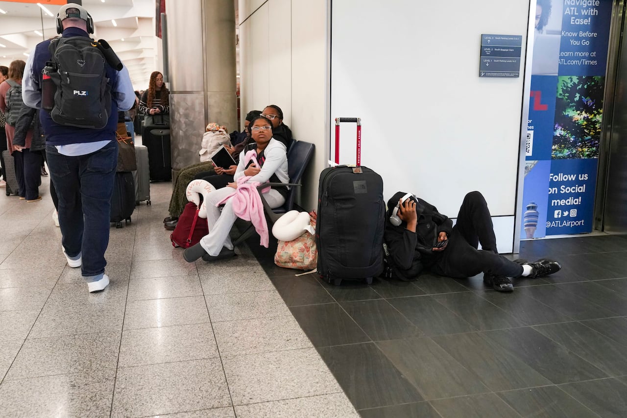 A young woman, wearing glasses and a white hoodie, sits on a chair in an airport surrounded by other people and baggage. To her right, a man lies on the floor next to a suitcase.