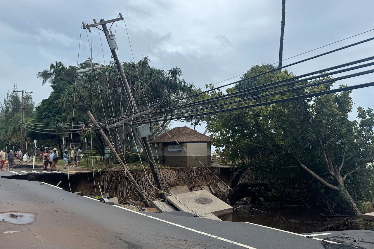 People in the background stand next to a washed out road with a tall utility pole and a large tree in a pit of water.