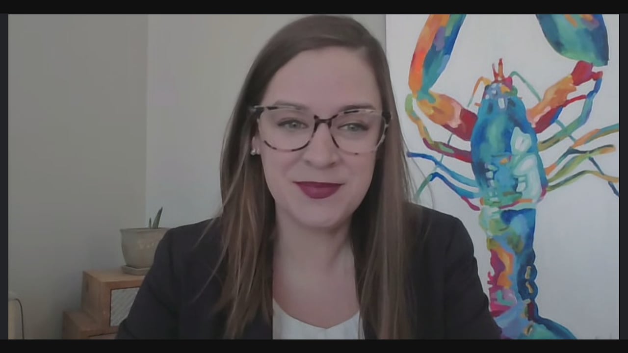 A woman in glasses and a dark top sits in front of a painting of a colourful lobster.