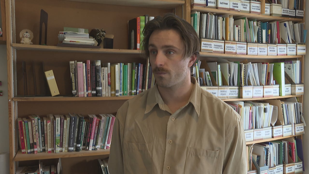 A man in a brown button-up shirt stands in front of shelves of books and files.