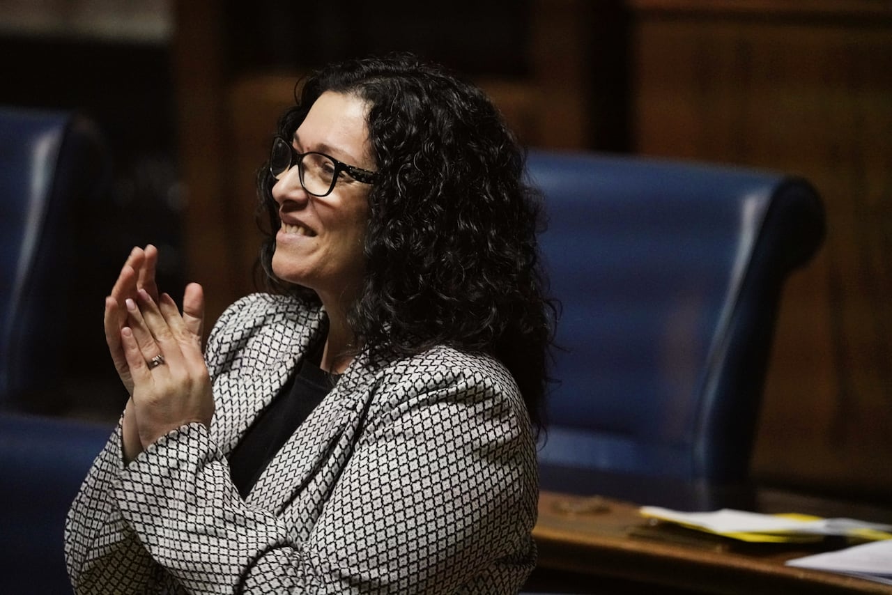 A woman in a black and white patterned jacket claps during a government legislative session.