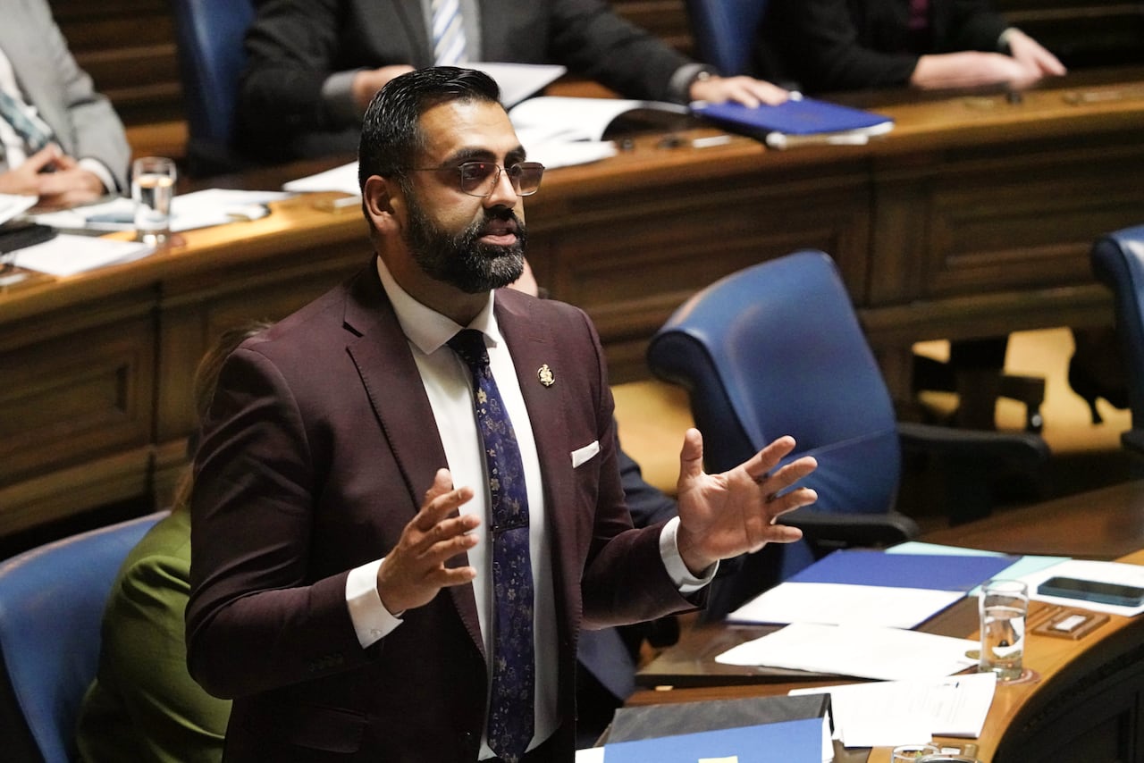 A man with short dark hair in a maroon suit gestures with his hands while speaking during a government legislative session.